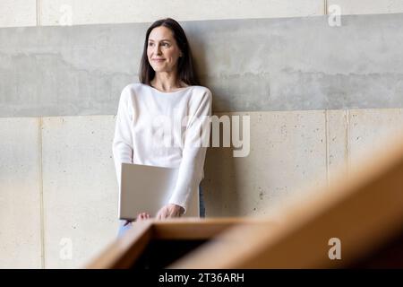 Femme d'affaires mûre réfléchie debout avec ordinateur portable devant le mur Banque D'Images