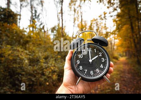 Gersthofen, Bavière, Allemagne. 23 octobre 2023. Main tenant le réveil noir avec 2 o horloge symbolisant l'heure d'hiver dans la forêt d'automne *** main hält einen schwarzen Wecker mit 2 Uhr als Symbol für Winterzeit in einem herbstlichen Wald crédit : Imago/Alamy Live News Banque D'Images