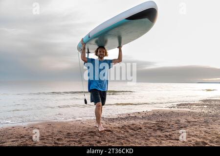 Homme souriant portant le paddleboard et marchant à la plage Banque D'Images