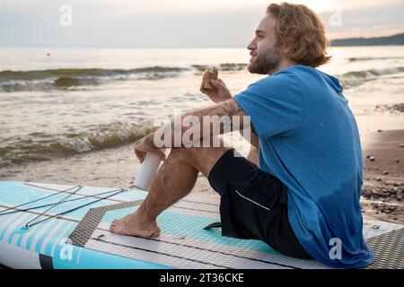 Homme blond mangeant un sandwich assis sur un paddleboard à la plage Banque D'Images