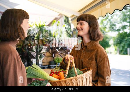 Femme souriante avec un panier de légumes parlant à sa sœur au marché fermier Banque D'Images