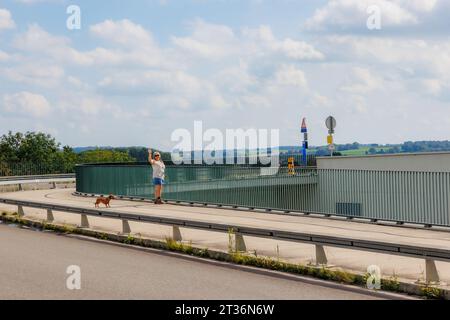 Route rurale et chemin piétonnier et vélos à Lanaye Locks, touriste senior souriante debout avec son chien à côté d'une clôture métallique agitant avec rai Banque D'Images