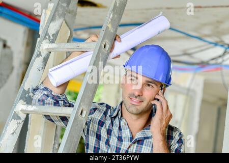 Constructeur au téléphone, appuyé sur l'échelle et tenant la paperasse enroulée Banque D'Images