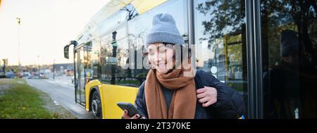 Image d'une jeune étudiante en attente des transports en commun, vérifie l'horaire sur l'application pour smartphone, se trouve près du bus de la ville Banque D'Images