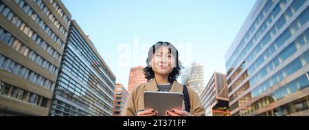 Portrait de sourires de fille coréenne heureux, marche en ville avec tablette numérique et sac à dos, utilise son gadget pour se déplacer Banque D'Images