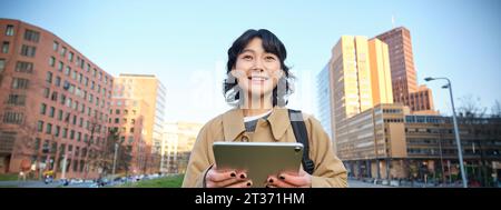 Une fille asiatique heureuse se tient dans la rue, un étudiant de l'université marche avec une tablette numérique dans les mains et les sourires, se tient dans le centre-ville Banque D'Images
