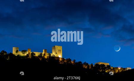 Château de Santa Catalina à Jaén, Espagne, illuminé la nuit, avec un ciel bleu nuageux, et le croissant de lune ciré sur le côté droit de l'image. Banque D'Images