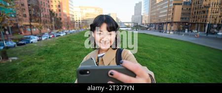 Jeune femme heureuse, étudiante prend le selfie dans la rue, tient sa tablette et ses notes de devoirs, utilise un smartphone Banque D'Images