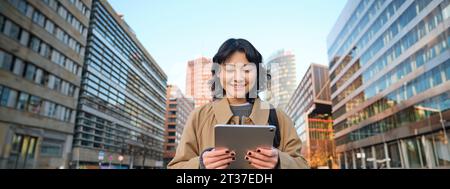 Portrait de sourires de fille coréenne heureux, marche en ville avec tablette numérique et sac à dos, utilise son gadget pour se déplacer Banque D'Images