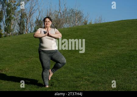 une femme d'âge moyen en surpoids pratique le yoga à l'extérieur pieds nus, faisant des exercices d'équilibre sur une jambe, pose d'arbre. Fitness, entraînement en plein air, yoga, poids l Banque D'Images