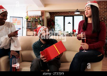 Homme caucasien joyeux et excité recevant une boîte cadeau festive de noël de la part de ses collègues. Employé de la société de démarrage assis sur le canapé dans le bureau et obtenir un cadeau de collègues Banque D'Images