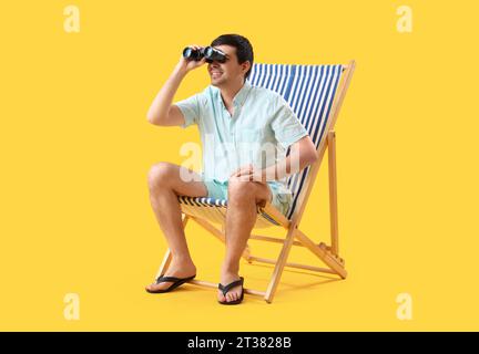 Jeune homme avec des jumelles assis dans une chaise longue sur fond jaune Banque D'Images