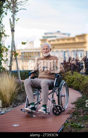 Homme âgé dans un fauteuil roulant assis à l'extérieur dans un jardin urbain, profitant du café et chaude journée d'automne. Portrait d'un homme âgé élégant aux cheveux gris Banque D'Images