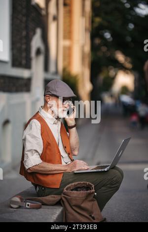 Homme âgé assis sur le trottoir de la rue travaillant sur un ordinateur portable, en utilisant les technologies numériques, faire un appel avec smartphone. Concept de seniors et compétences numériques. Banque D'Images