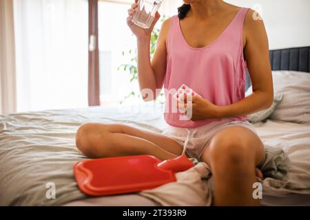 Femme à la maison souffrant de douleurs menstruelles, prenant des analgésiques. Crampes menstruelles, femme réchauffant le bas-ventre avec une bouteille d'eau chaude, endométriose Banque D'Images