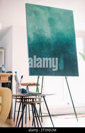 Design créatif de la chambre avec peinture à l'aquarelle vert mer placée sur chevalet contre le mur blanc dans un studio moderne confortable pendant près de la table en bois à la maison Banque D'Images