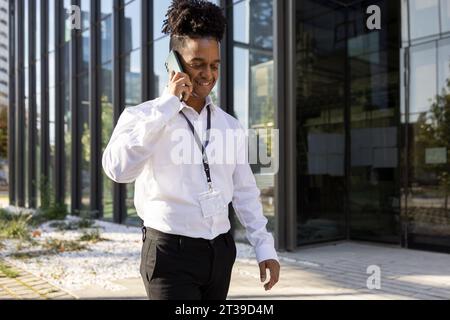 Portrait de joyeux entrepreneur afro-américain en tenue formelle parlant sur smartphone tout en marchant dans la rue de la ville Banque D'Images