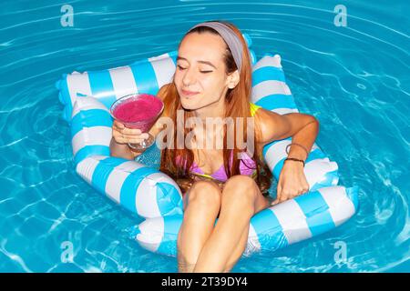Jeune femme se détendant sur un radeau gonflable dans une piscine, tenant une boisson de cocktail rose, profitant de sa journée d'été. Banque D'Images