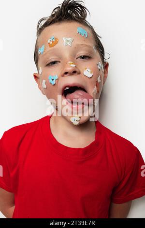 Un jeune garçon avec les cheveux mouillés sort de sa langue et a des autocollants animaux colorés sur son visage, portant une chemise rouge sur un fond blanc. Banque D'Images