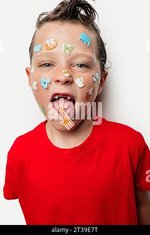 Un jeune garçon avec les cheveux mouillés sort de sa langue et a des autocollants animaux colorés sur son visage, portant une chemise rouge sur un fond blanc. Banque D'Images