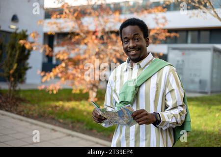 Portrait d'homme afro-américain souriant regardant la caméra tout en se tenant sur une route pavée avec une carte contre des arbres complexes de bâtiment brouillés à la lumière du jour Banque D'Images