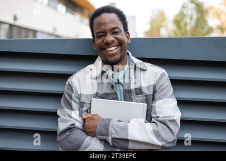 Portrait d'homme afro-américain souriant drôle regardant la caméra tout en portant un ordinateur portable dans les bras croisés contre le bâtiment flou Banque D'Images
