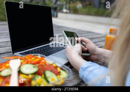 Vue rapprochée de jeune femme étudiante méconnaissable en temps de pause et communique en ligne sur le téléphone avec un ordinateur portable et une salade près de son outsidd Banque D'Images