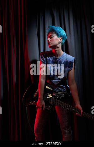 Jeune musicienne sérieuse dans les cheveux teints et tissu décontracté avec la guitare portant dans les mains debout tout en jouant sur scène Banque D'Images