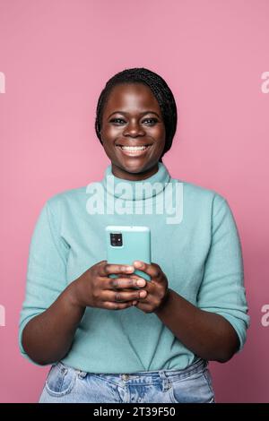 Une jeune femme joyeuse dans un col roulé turquoise tient un smartphone bleu clair, souriant largement sur un fond rose Banque D'Images