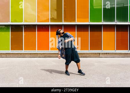 Un homme latin avec des cheveux tressés, des lunettes de soleil et des écouteurs se penche contre un mur de verre coloré, portant une chemise florale bleue, un pantalon noir et un snea Banque D'Images