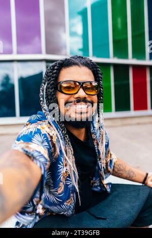 Portrait rapproché d'un homme latin souriant avec des tresses et des lunettes de soleil, avec des structures en verre colorées en arrière-plan. Banque D'Images