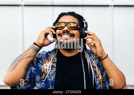 Homme latin avec des tresses et des lunettes de soleil appréciant la musique dans un casque noir sur un fond urbain blanc. Banque D'Images