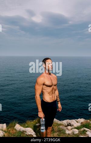 Un homme avec un physique tonique se tient près de la mer tenant une bouteille d'eau. Banque D'Images