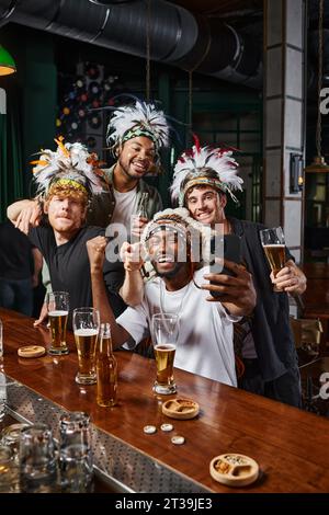 joyeux hommes multiethniques en coiffe avec des plumes prenant selfie sur smartphone pendant la fête de célibataire Banque D'Images