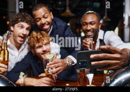 collègues multiethniques joyeux en tenue formelle prenant selfie sur smartphone dans le bar après le travail Banque D'Images