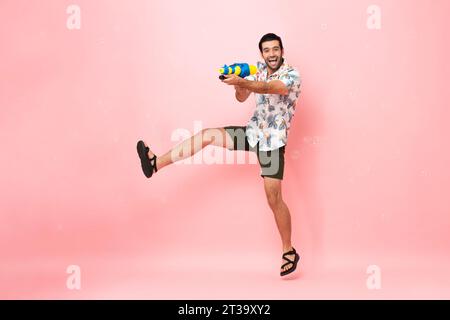 Beau souriant jeune homme touristique caucasien jouant avec un pistolet à eau et sautant dans le fond rose studio pour le festival Songkran en Thaïlande et southe Banque D'Images