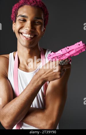 portrait de jeune homme joyeux avec les cheveux roses et les bretelles posant pistolet jouet et souriant joyeusement Banque D'Images