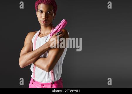 modèle masculin beau à la mode avec les cheveux roses posant avec pistolet jouet regardant sérieusement la caméra Banque D'Images