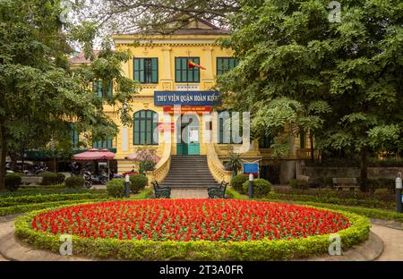 Le grand bâtiment colonial français abritant la bibliothèque du district de Hoan Kiem avec des lits de fleurs ornés dans le parc en face, Hanoi, Vietnam. Banque D'Images