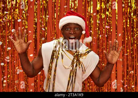 homme afro-américain émerveillé en chapeau de père noël et débardeur blanc montrant le geste wow sur fond brillant Banque D'Images