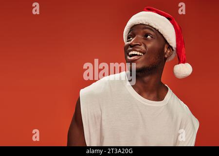 homme afro-américain étonné et débordé de joie dans le chapeau du père noël souriant et regardant loin sur fond rouge Banque D'Images