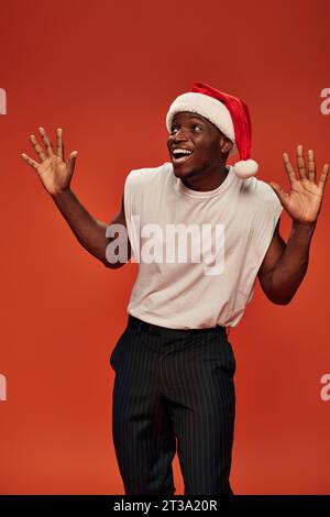 homme afro-américain étonné et débordé de joie dans le chapeau de santa montrant le geste wow et regardant loin sur le rouge Banque D'Images