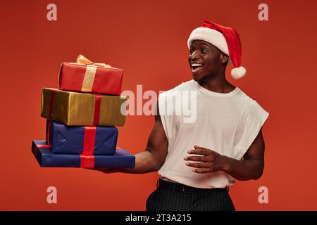 homme afro-américain étonné et débordé de joie dans le chapeau de santa regardant des boîtes cadeaux colorées sur rouge Banque D'Images