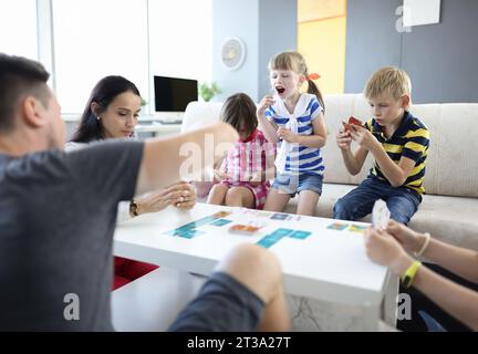 Les adultes et les enfants sont assis à la table et tiennent les cartes de jeu que la fille se leva et crie. Banque D'Images