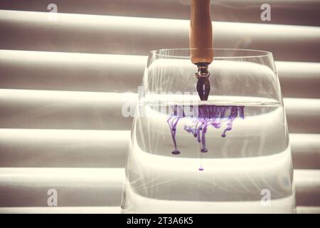 pointe métallique de stylo à encre trempée dans l'eau. diffusion de l'encre bleue dans une tasse en verre transparent avec de l'eau claire sur le fond d'un écran rayé Banque D'Images