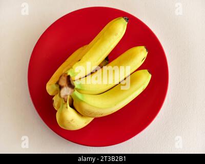 Bouquet de bananes fraîches sur une assiette rouge sur la table blanche. Vue de dessus. Banque D'Images