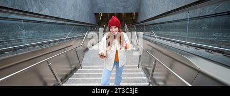 Photo en extérieur d'une jeune femme qui planifie un itinéraire, suit la carte sur l'application pour smartphone, monte les escaliers avec un sac à dos et sourit Banque D'Images