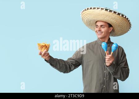 Beau jeune homme en sombrero avec maracas et bol de nachos sur fond bleu Banque D'Images