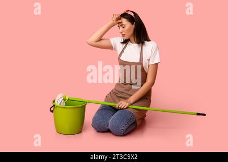 Portrait de jeune femme au foyer fatiguée en tablier avec vadrouille et seau sur fond rose Banque D'Images