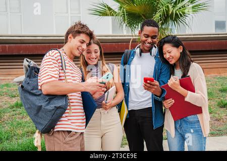 Heureux camarades de classe divers regardant leurs smartphones et riant ensemble en regardant des vidéos drôles après la classe. Jeunes étudiants multiraciaux utilisant leur téléphone cellulaire et partageant du contenu sur les réseaux sociaux. Photo de haute qualité Banque D'Images
